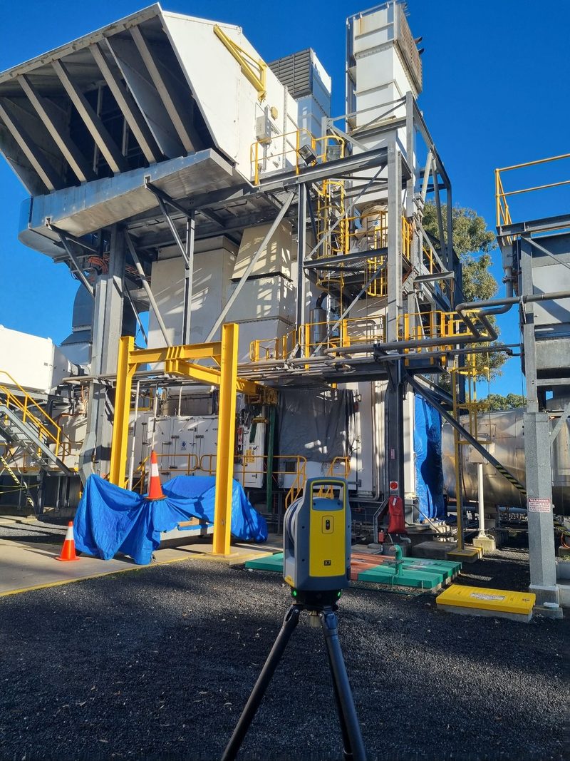 Trimble X7 laser scanner on a tripod at ground level beside an industrial plant structure with steel stairs and platforms