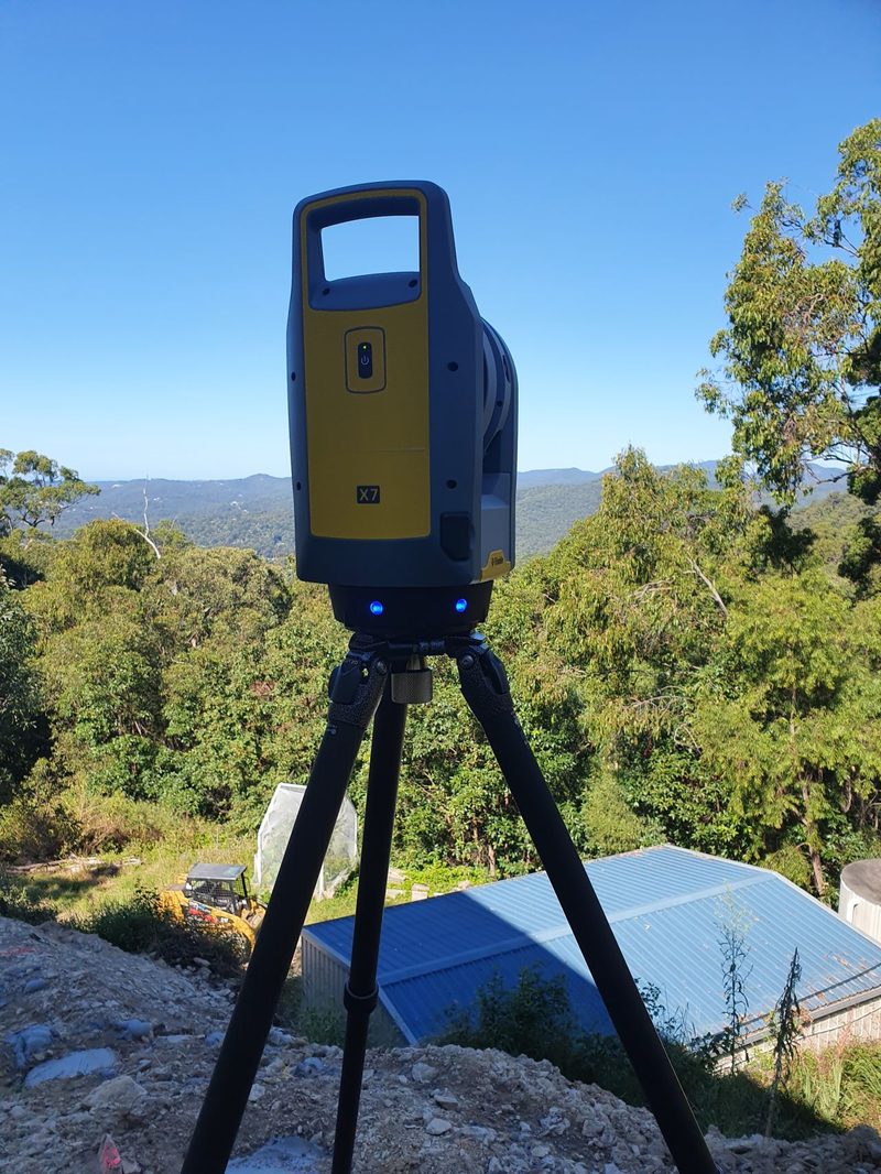 Trimble X7 laser scanner on a tripod on a hillside above a building roof, with bushland behind